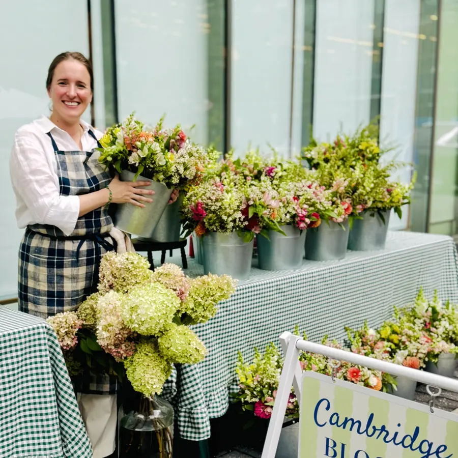 Alba in a plaid apron holding a bucket of flowers beside the Cambridge Blooms sign